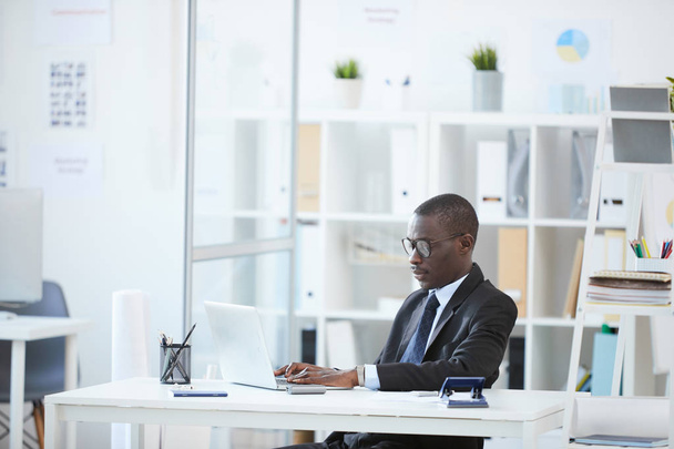 Skoncentrowany afrykański biznesmen w okularach wpisujący na klawiaturze laptopa siedząc w biurze w miejscu pracy - Zdjęcie, obraz