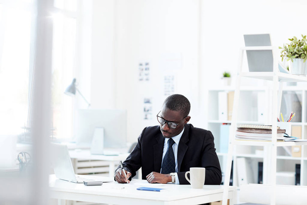 Poważny afrykański biznesmen w okularach siedzi przy stole i pisze coś w biurze - Zdjęcie, obraz