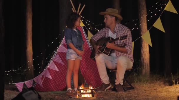 loving father plays guitar for his smiling daughter while camping in woods near fire on background of wigwam - 映像、動画