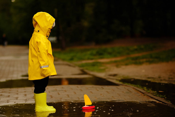 黄色いレインコートを着た少年は、水たまりに浮かぶおもちゃの船を見る。公園を歩いている小さな赤ちゃん. - 写真・画像