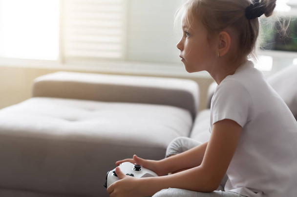 Enthusiastic kid in casual outfit playing with joystick while sitting on sofa in living-room - Foto, immagini