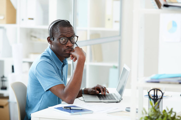 Портрет африканського дизайнера у навушниках і окулярах, що сидять на робочому місці перед ноутбуком і дивляться на камеру в офісі. - Фото, зображення