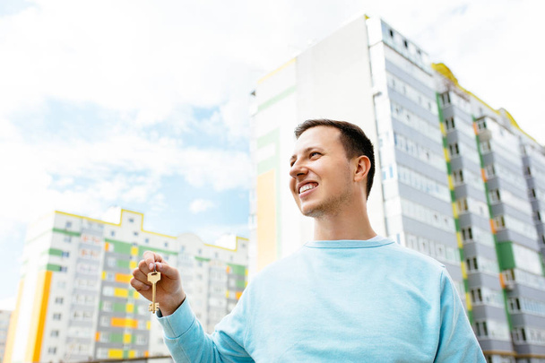 Молодой человек стоит с ключами от новой квартиры в руке на фоне нового многоэтажного дома
 - Фото, изображение