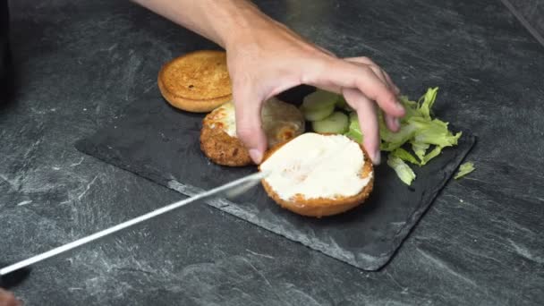 Αρσενικό χέρι σε τατουάζ που διαδίδει τυρί κρέμα σε ένα σάντουιτς και βάζοντας μαρούλι, αγγούρι και κρέας - Πλάνα, βίντεο