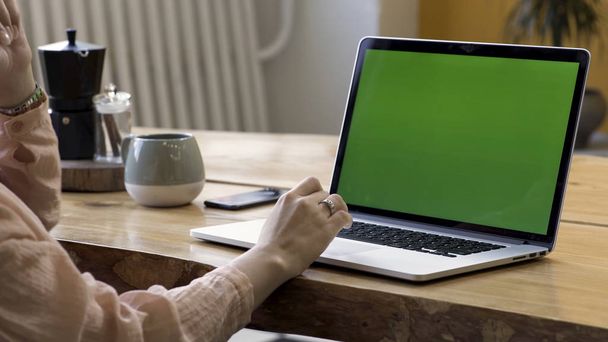 近代的なアパートのテーブルの上に立っている緑色の画面を持つ彼女のラップトップを使用して、指に金の指輪を持つ女性の手のためにクローズアップ。ストック映像。自宅での職場、クロマキー付きラップトップ. - 写真・画像