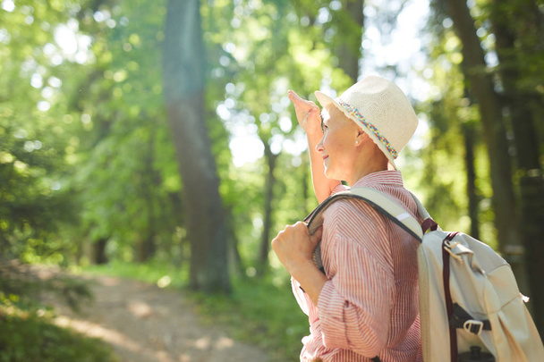 Portrait of casual senior woman in hat and with backpack smiling while standing outdoors in sunny summer - Foto, Imagen