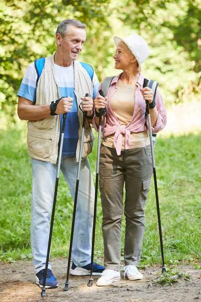 Portrait of mature woman in hat and with backpack looking at camera while doing nordic walking together with husband in the nature - Zdjęcie, obraz