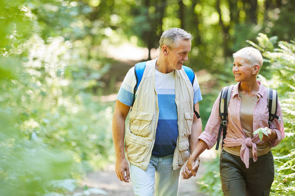 Lovely mature couple with backpacks holding hands and talking to each other during walk in the forest in sunny summer - Foto, immagini