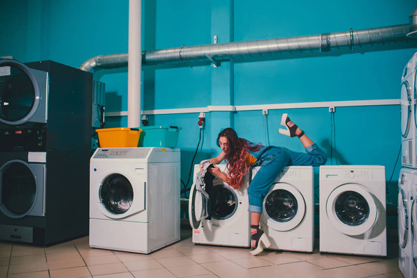 Mujer joven buscando ropa en la lavadora tambor en la lavandería
. - Foto, Imagen