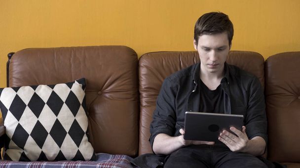 Egy feszült, fekete inges férfi használja a tabletjét, miközben otthon ül a barna bőrkanapén. Készletfelvétel. Stresszes hipster blogger öltözött fekete gazdaság modern touch pad eszköz. - Fotó, kép