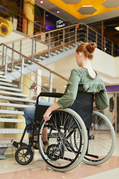 Mujer pelirroja sentada en silla de ruedas mirando escaleras pensando cómo subir escaleras en la oficina
 - Foto, Imagen