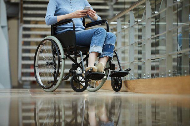 Κοντινό πλάνο νεαρής ανάπηρης γυναίκας με περιστασιακή ενδυμασία που κάθεται σε αναπηρική καρέκλα και πληκτρολογεί μήνυμα στο κινητό τηλέφωνο - Φωτογραφία, εικόνα