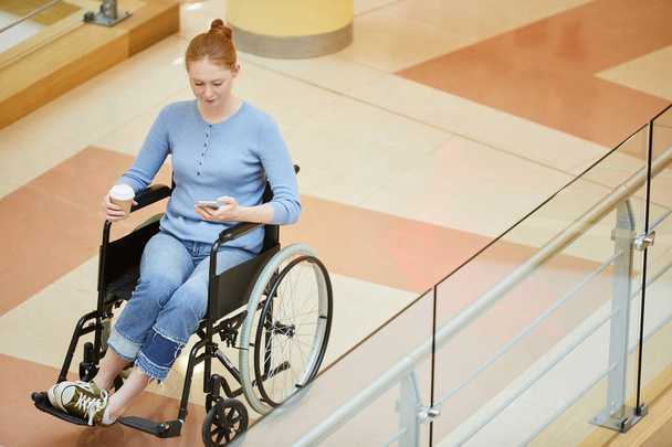 Jovem mulher com deficiência em roupas casuais sentada em cadeira de rodas e olhando para o telefone celular enquanto bebe café de copo descartável
 - Foto, Imagem