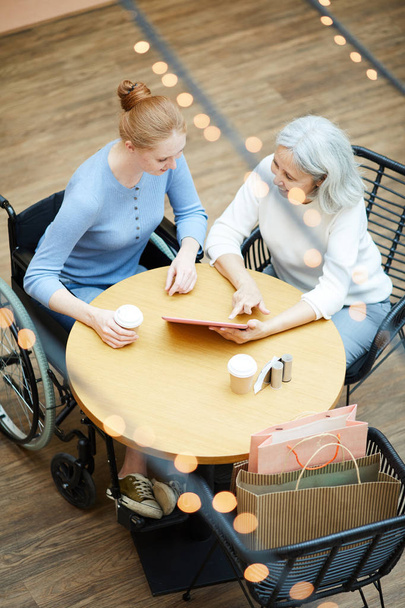 junge behinderte Frau zeigt auf digitales Tablet und erklärt Seniorin etwas, während sie im Café irgendein Dokument ausfüllt - Foto, Bild