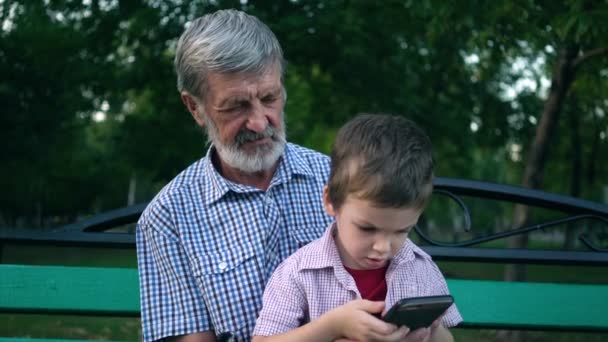 Senior grandfather and grandson are sitting on a bench in the park and playing on a smartphone - Footage, Video