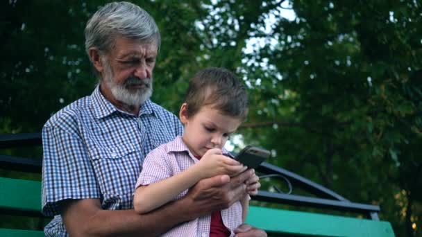 Старший дедушка и внук сидят на скамейке в парке и играют на смартфоне
 - Кадры, видео