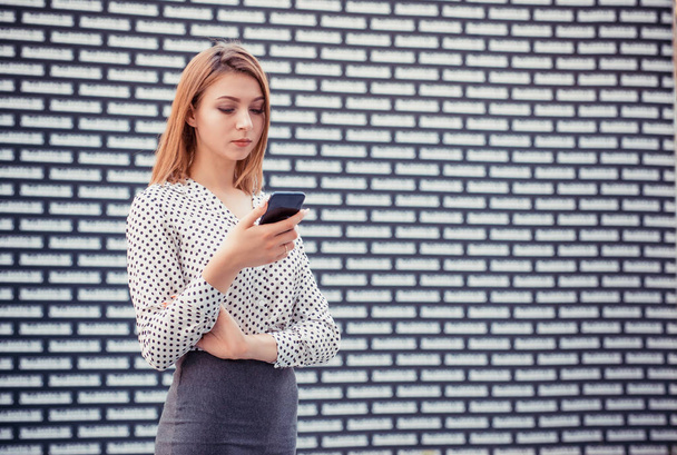 Portrait of beautiful young woman surfing the Internet on cell phone, looking at screen with concentrated expression, checking email, girl wearing formal black suit, shirt on white black bricks wal - Foto, Imagem