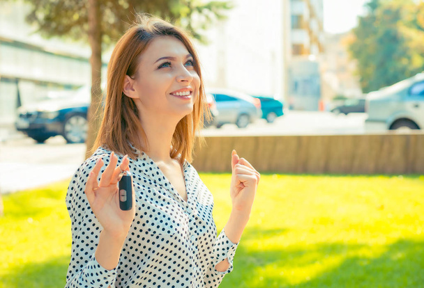 Just won a car!!! Super excited smiling laughing brunette girl woman celebrating driver holding key of her new car automobile outside in summer day in city building and green trees on background - 写真・画像