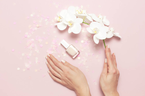 Woman's hands with gentle manicure hold branch of White Phalaenopsis orchid flowers on pastel pink background with festive confetti top view flat lay. Tropical flower. Holiday Women's Day Flower Card - Photo, Image