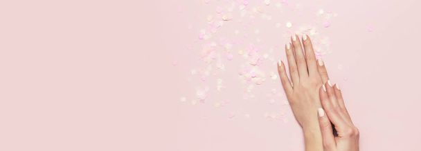 Stylish fashionable female manicure in pastel colors. Hands of young girl on pink background with festive confetti. Minimalist manicure trend. Flat lay, top view, copy space for your text - 写真・画像