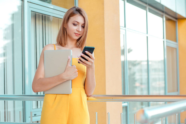 Charming young woman, student in casual yellow jumpsuit looking at mobile phone and holding computer in other hand standing outdoors office, college, university building on background - Zdjęcie, obraz