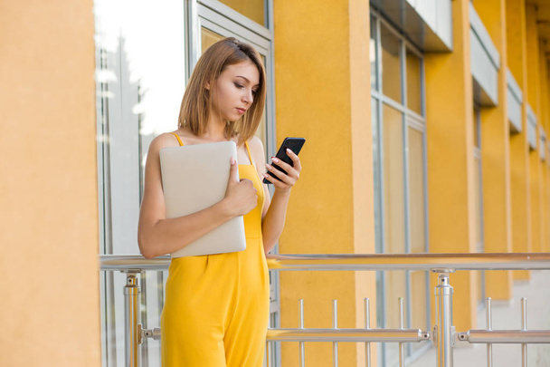 Charming young woman, student in casual yellow jumpsuit looking at mobile phone and holding computer in other hand standing outdoors office, college, university building on background - Photo, Image