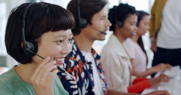 Side view close up of a smiling young Asian female call centre worker typing and talking on a phone headset in a modern open plan office, with a group of young multi-ethnic colleagues wearing headsets and using computers working in the background - Metraje, vídeo