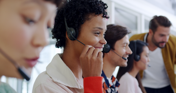 Side view close up of a group of young multi-ethnic call centre workers wearing headsets and using computers in a modern open plan office, with a young Caucasian male manager overseeing their work in the background - Video