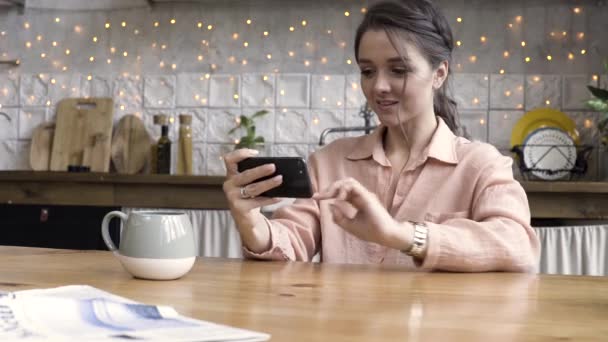 Young womanplaying smartphone games at the kitchen table with coffee mug in a modern home. Stock footage. Beautiful brunette female looking at her smartphone on kitchen stuff background. - Filmati, video