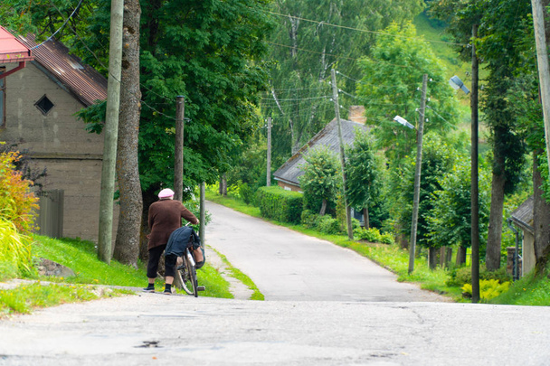 Rückansicht einer alten Frau, die im Sommer ein Fahrrad auf einer kleinen Stadtstraße schiebt - Foto, Bild