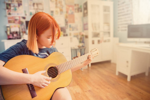 Pretty red-haired teenage girl plays guitar in her room - Photo, Image