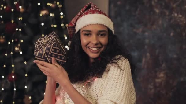 Young Pretty Mulatto Woman at Santas Hat and Decorative Lights on her Neck Shaking Present Box in her Hands, Looking to Camera and Smiling at Cosy Home Background. Holiday Celebration Concept. - Séquence, vidéo