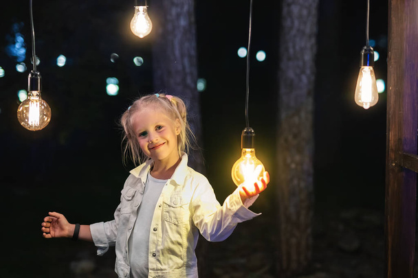Мила чарівна біла дівчина портрет посміхається і тримає в руці одну з повішених лампочок на відкритому повітрі. Правильна концепція вибору рішення
 - Фото, зображення