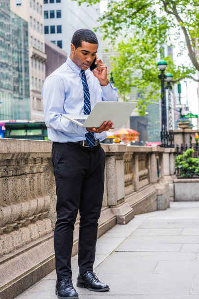 Giovane uomo d'affari afroamericano che lavora al computer portatile, parla al telefono, sta in piedi davanti a mezzo muro di marmo in strada a Manhattan, New York, indossa camicia bianca, cravatta, pantaloni neri, scarpe di pelle
 - Foto, immagini