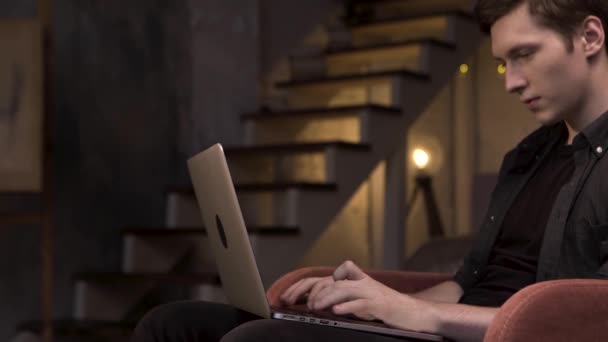 Side view of young man using laptop while sitting at home on stairs background. Stock footage. Man sitting in the red armchair and typing on his computer in the dim room. - Felvétel, videó