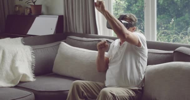 Front view of a senior Caucasian man wearing a VR headset and reaching with his hands out and boxing, sitting on a sofa - Imágenes, Vídeo