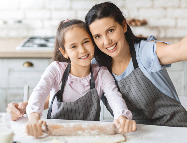 Χαριτωμένο μικρό κορίτσι και την όμορφη μαμά της σε ποδιές παίρνοντας selfie, χαμογελώντας στην κουζίνα - Φωτογραφία, εικόνα