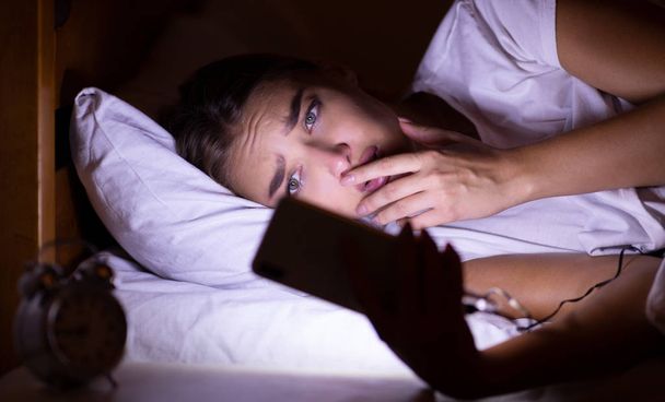 Ανήσυχος κορίτσι χρησιμοποιώντας το κινητό τηλέφωνο ξαπλωμένος στο κρεβάτι τη νύχτα - Φωτογραφία, εικόνα