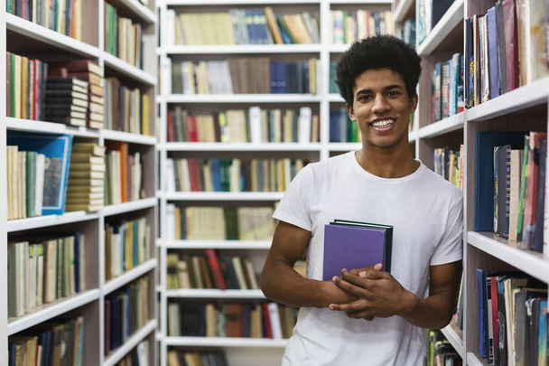 Cheerful african american guy staying between bookshelves, holding book - Photo, Image