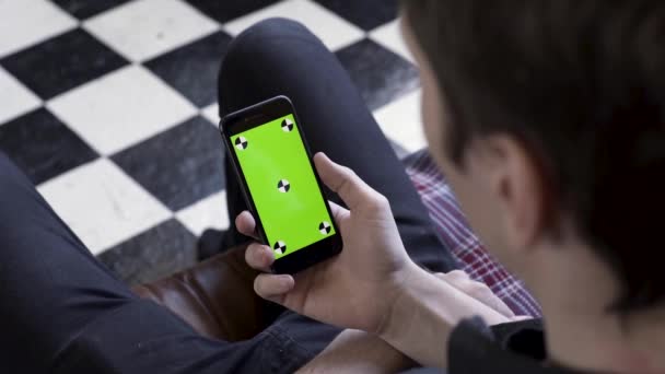 Přes rameno pohled na muže pomocí chytrého telefonu se zelenou obrazovkou v místnosti s šachovnicovou podlahou. Záběry ze skladu. Muž drží své zařízení s chromakey clonou a dívá se na něj. - Záběry, video