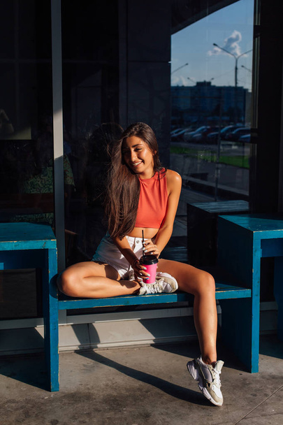 Stylish happy young brunette woman wearing white shorts and sneakers holding pink cup of coffee to go. - Fotografie, Obrázek