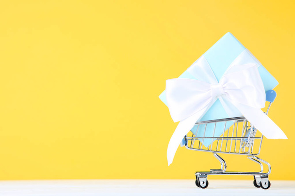 Small shopping cart with gift box on yellow background - 写真・画像