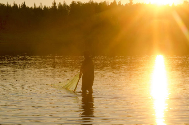Um homem solitário, um pescador Yakut em botas vadias wearily vai para a rede para a vida selvagem no rio Vilyuy nos raios brilhantes do sol pegando o chugunok peixe local ao pôr do sol
. - Foto, Imagem