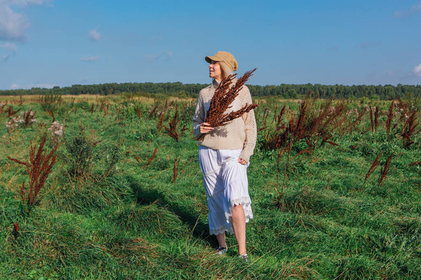 Happy beautiful blond woman walking in a green field with a bouquete of dry brown plants - Fotografie, Obrázek