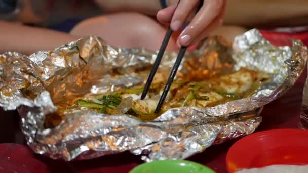 Turisté jedí ryby pomocí hůlek vařených na fólie na trhu s potravinami v asijském ulicích. Detailní - Záběry, video