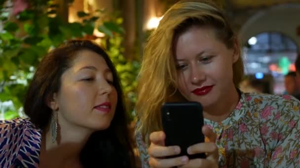 Δύο νεαρές γυναίκες, που κάθονται σε ένα εστιατόριο και περιμένουν τα πιάτα τους, επιλεγμένα για δείπνο. Οι φίλοι διασκεδάζουν βλέποντας φωτογραφίες στο smartphone. Δύο χαρούμενες γυναίκες που μιλούν και κοιτάζουν σε ένα - Πλάνα, βίντεο