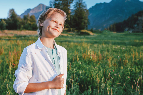 Rubia sonriente chico en el fondo de la maravillosa vista de la pradera verde y las montañas, familia viaje aventura estilo de vida
 - Foto, Imagen