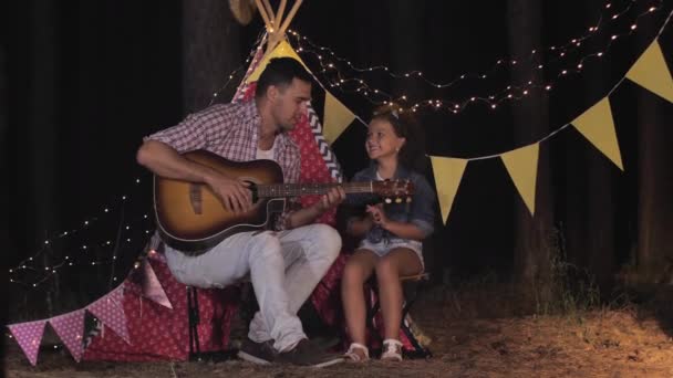 relaxing with parent, beautiful curly girl has fun talking with her father and playing guitar during forest party on family weekend on background of wigwam - 映像、動画