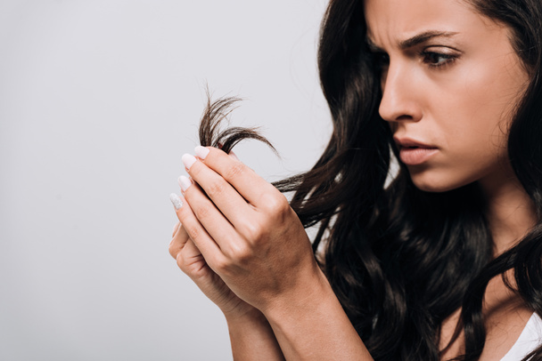rozčilená brunetka krásná žena, která hledí na poškozené vlasy izolované na šedé - Fotografie, Obrázek
