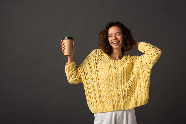 heureuse femme bouclée en chandail tricoté jaune avec les yeux fermés tenant du café pour aller sur fond noir
 - Photo, image
